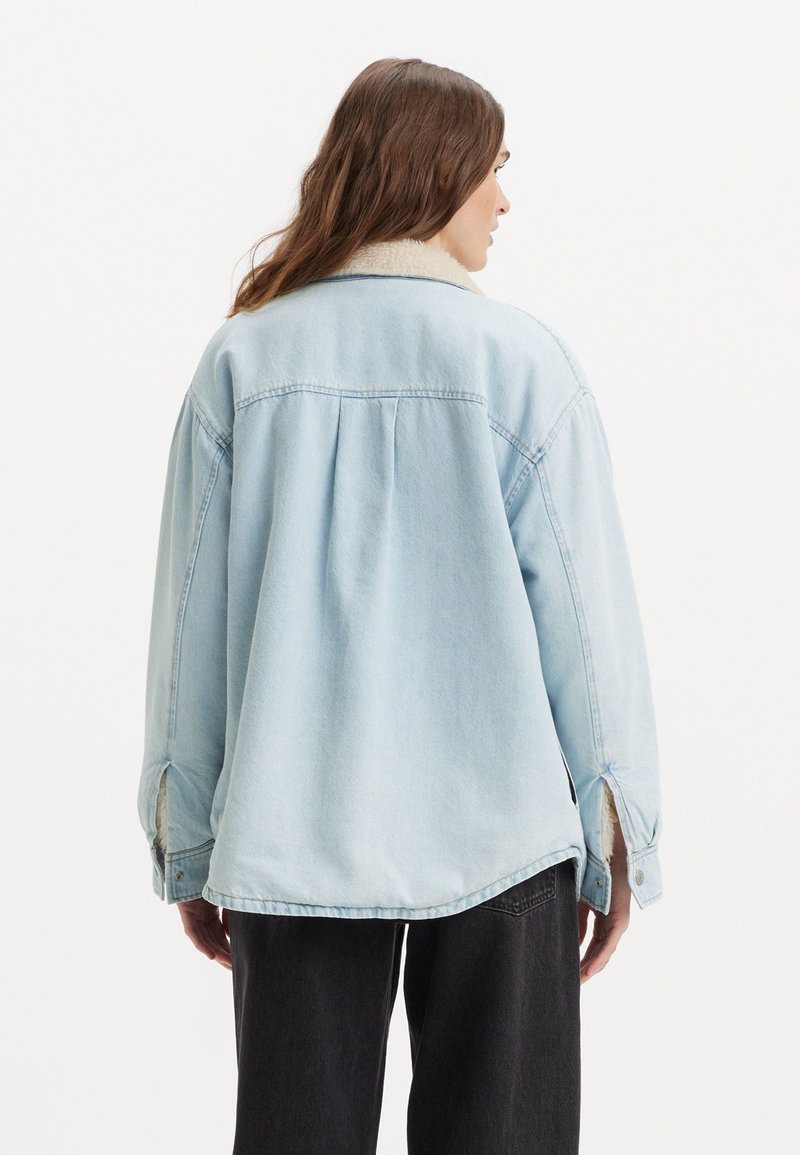 Chaqueta de denim azul claro con un acogedor cuello de borreguillo, hombros caídos y un ajuste casual y holgado, con aberturas en los laterales del dobladillo.