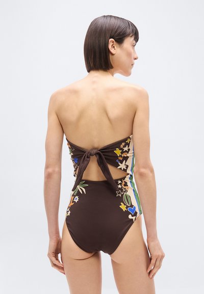 Γυναίκα με κοντά σκούρα μαλλιά, ντυμένη με καφέ ολόσωμο μαγιό με λουλουδάτα σχέδια και δέσιμο φιόγκος στο πίσω μέρος, στέκεται μπροστά σε ένα απλό φόντο.