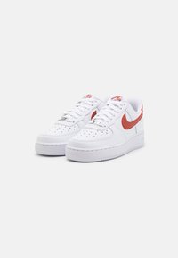 Fehér bőr sportcipők piros swoosh-sal és díszítésekkel, perforált orr-résszel, lapos fűzőkkel és texturált gumitalppal.