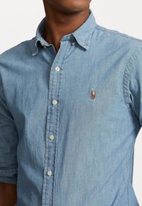 Camicia in chambray azzurro chiaro con maniche corte, colletto button-down, bottoni visibili e logo ricamato sul petto. Tessuto morbido.