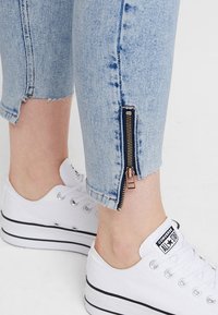 Jeans cropped in denim azzurro chiaro con dettaglio zip lungo la cucitura esterna e orlo sfrangiato, abbinati a sneaker bianche Converse.