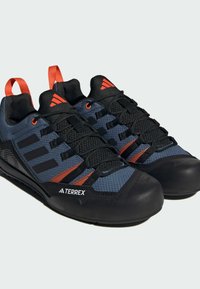 Niebieskie i czarne buty do wspinaczki z czerwonymi akcentami, teksturowana siateczkowa cholewka i czarne gumowe podeszwy. Oferują mocne sznurowanie oraz ikoniczne logo z trzema paskami.