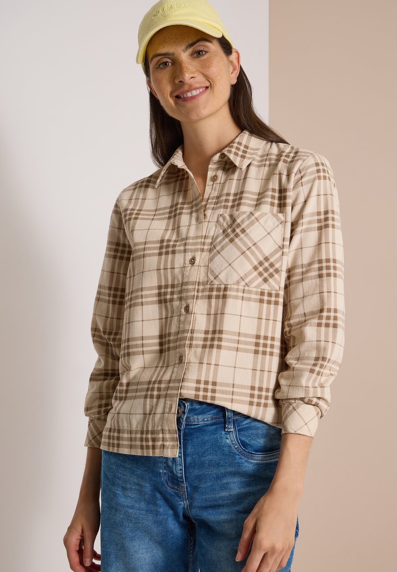 Femme portant une chemise à carreaux beige et marron boutonnée, un jean bleu et une casquette jaune clair, souriant à la caméra sur un fond bicolore.