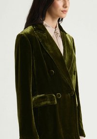 Blazer croisé en velours vert riche. Comprend des revers crantés, deux gros boutons et une poche latérale. Texture lisse et coupe ajustée.