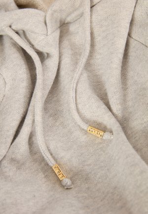 Haut à capuche gris clair avec une texture douce, des cordons de serrage avec des embouts en métal doré et une coupe décontractée. Des détails de couture élaborés sont visibles.