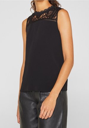 Vrouw draagt een zwart mouwloos topje met kantdetail bij het beleg en zwarte leren broek, staand tegen een effen achtergrond.