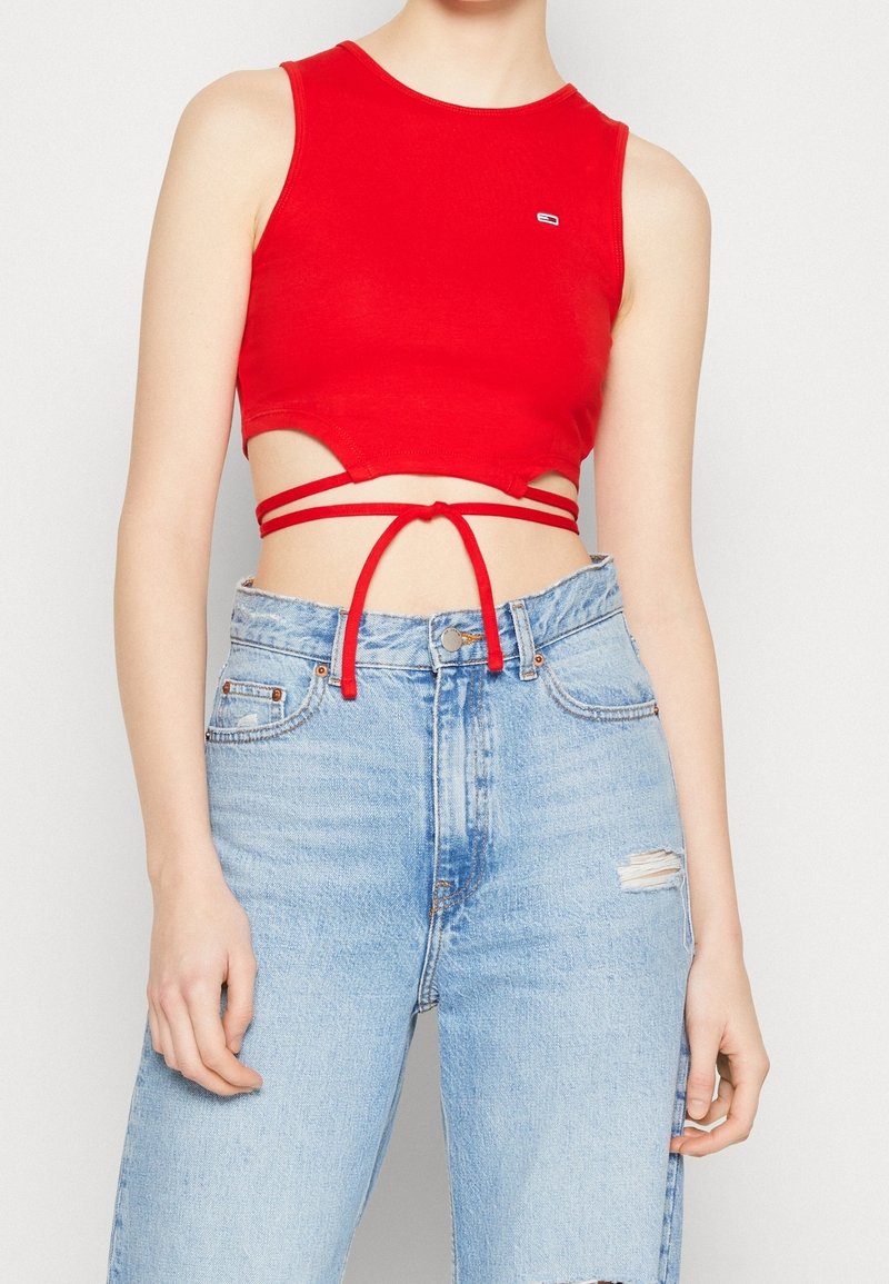 Top corto rojo con diseño sin mangas, cintura recortada y detalles de atado, combinado con jeans descoloridos azul claro. Material de algodón suave.