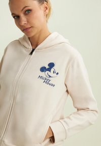 Sweat à zip couleur beige clair avec un imprimé Mickey Mouse sur le devant en bleu foncé. Comprend une capuche doublée et une texture lisse et douce.
