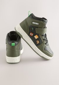 Sneakers alte in pelle verde con dettagli neri, suola bianca macchiata e patch a tema Minecraft con piccone e TNT.