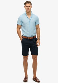Jasnoniebieska, krótko rękawowa koszula z guzikami, granatowe szorty, brązowy skórzany pasek i brązowe mokasyny. Prosty, codzienny strój na ciepłą pogodę.