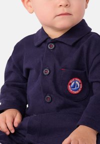 Mørkeblå barne skjorte laget av ribbestrikket stoff med krage, med sirkulære knapper og et brodert logo-patch på brystet.