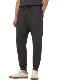 Muž v černých joggingových kalhotách s elastickými lemy a bílých teniskách, stojící s jednou rukou v kapse na bílém pozadí.