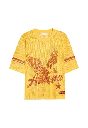 Jeune homme avec des dreadlocks portant un t-shirt en maille jaune avec un motif d'aigle et une chemise à carreaux nouée autour de sa taille, debout près d'un buste en pierre et d'un mur en briques.