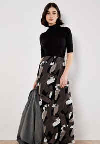 Femme avec les cheveux courts et foncés portant un col roulé noir et une longue jupe à motifs abstraits noir, gris et blanc, tenant un manteau gris.