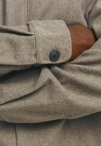 Manica di camicia in tessuto a spina di pesce marrone con pattina, dotata di un bottone nero e dettagli di cucitura visibili. Texture morbida.