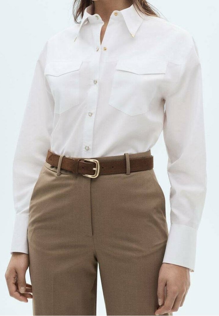 Chemise blanche à manches longues avec poches poitrine, boutons dorés en accent, associée à une ceinture marron et un pantalon taille haute beige. Texture lisse.