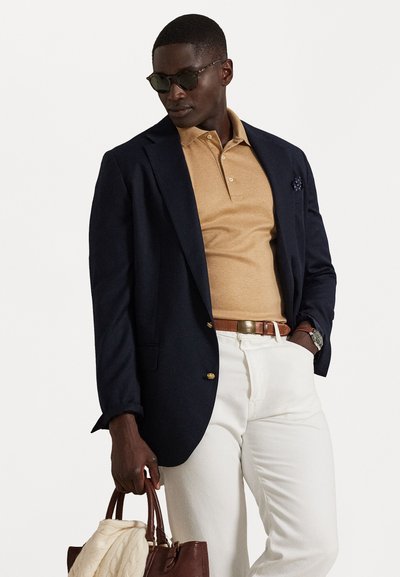 Ναυτικό σακάκι, μπεζ πόλο, λευκά παντελόνια, καφέ ζώνη και καφέ δερμάτινη τσάντα. Γυαλιά ηλίου και μαντήλι τσέπης προσθέτουν λεπτομέρεια στο ντύσιμο.