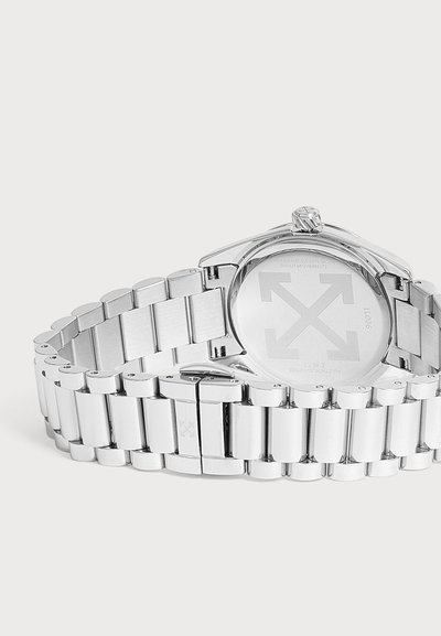 Montre bracelet en métal argenté avec bracelet à maillons vue de dos, affichant une gravure et le logo de la marque sur le boîtier et la boucle.