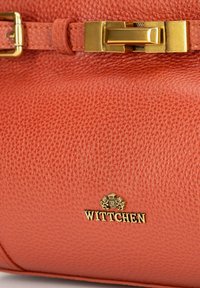 Terrakotowa skórzana torebka o chropowatej fakturze, z zapięciem na złotą klamrę i złotą metką z logo, która dodaje eleganckiego akcentu.