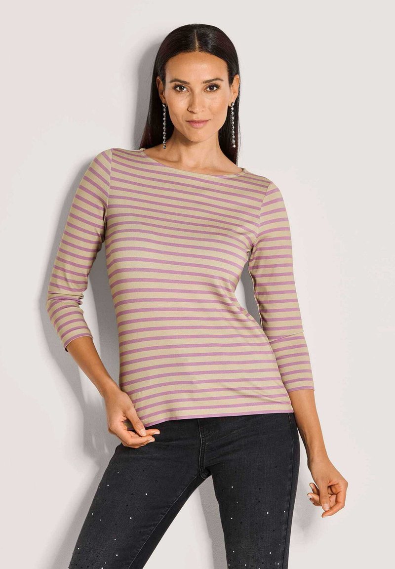 Vrouw in een paars en beige gestreepte driekwart mouwen top met zwarte jeans versierd met kleine witte stippen, poseert met één hand op de heup.