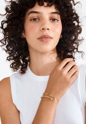 Žena s kovrčavom tamnom kosom, u bijeloj majici bez rukava i zlatnim narukvicama od lanca, nježno dodiruje svoje rame na bijeloj pozadini.