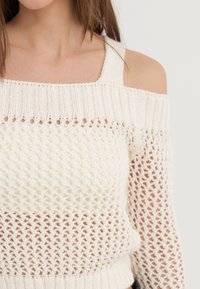 Sweter dzianinowy bez ramion w kolorze off-white, z teksturowanym wzorem i otwartymi ściegami oraz ściągaczem na dole. Lekka i oddychająca konstrukcja.