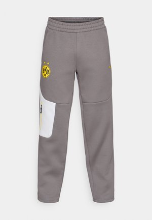 Pantalons de survêtement gris avec un patch blanc, taille élastique, logo jaune et accent zippé jaune. Texture douce et coupe fuselée.