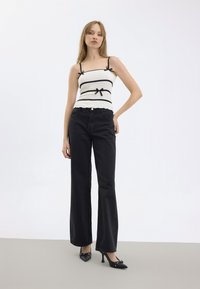 Schwarze hochgeschnittene ausgestellte Jeans, kombiniert mit einem weißen gerippten Oberteil mit schwarzen Streifen und Schleifen an den Trägern. Schwarze High Heels.