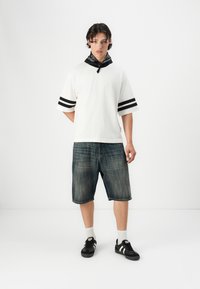 Hvid kortærmet skjorte med sorte striber, parret med knæ-lange blå denimshorts og sorte sko. Sort bandana bundet om halsen.