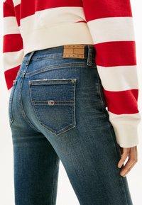 Jeans dżinsowe w ciemnym odcieniu, z wysokim stanem i aksamitną teksturą. Posiada skórzaną naszywkę z logo oraz mały haftowany detal flagi na tylnej kieszeni.