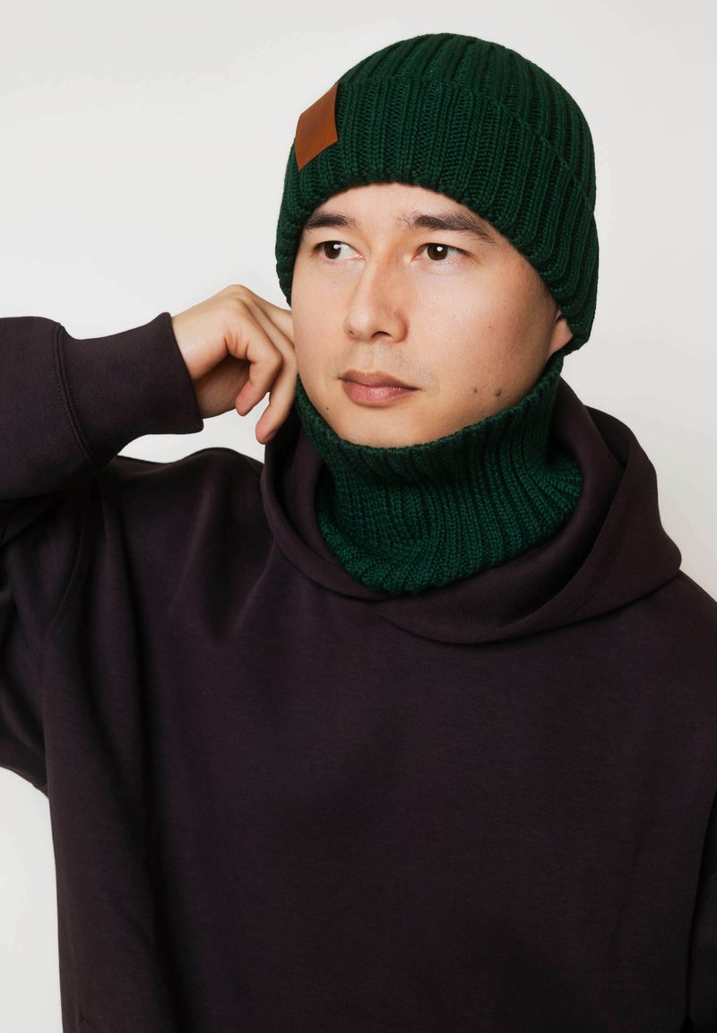 Ciemnozielona dzianinowa czapka z naszywką ze skóry, noszona razem z dopasowanym ciemnozielonym prążkowanym kominem, zestawiona z ciemnofioletową bluzą z kapturem.