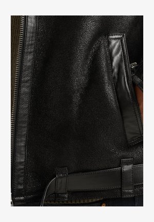 Detailný záber na čiernu textúrovanú koženú bundu, ktorý zobrazuje zips, vrecko a detail opaska s náznakom hnedej tkaniny pod ním.