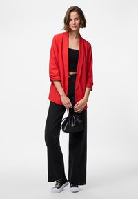Červený blazer s ohrnutými rukávy přes černý crop top, spárovaný s černými širokými kalhotami a černými teniskami, držící malou černou kabelku.