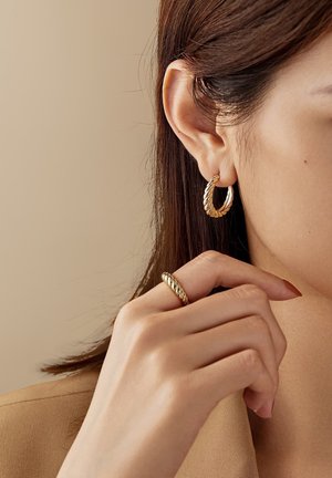 Femme aux cheveux bruns raides portant une boucle d'oreille en or torsadé et une bague assortie en or torsadé à un doigt près du visage.