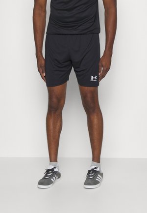 Mann trägt schwarze Under Armour Shorts, schwarzes Oberteil, graue Socken und graue Adidas-Sneaker und steht vor weißem Hintergrund.