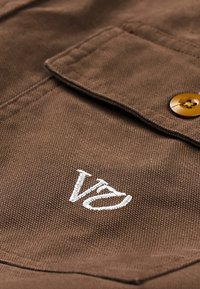 Smeđa tkanina s teksturiranim završetkom, s bijelim izvezenim logotipom i smeđim gumbom na džepu s preklopom.