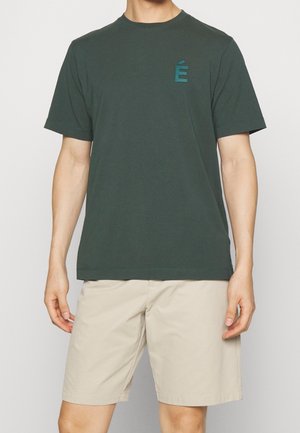 Dunkelgrünes Baumwoll-T-Shirt mit rundem Halsausschnitt und kurzen Ärmeln, das auf der linken Brusttasche einen tealfarbenen gestickten Buchstaben "É" zeigt. Kombiniert mit beigen Shorts.