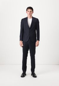 Mies seisoo yllään tummansiniraidallinen pukupuku, jossa on kaksinappinen bleiseri, valkoinen t-paita ja mustat juhlakengät valkoista studiotaustaa vasten.