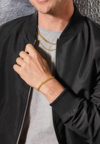 Uomo che indossa una giacca nera, una camicia grigia, una collana dorata e un braccialetto dorato, mentre tiene la zip della giacca vicino al colletto.
