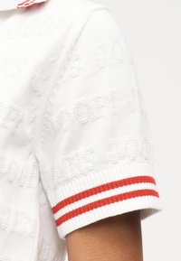 Λευκό πλεκτό τοπ με κοντά μανίκια, με διακριτικό ανάγλυφο κείμενο και ριμπ μανσέτες που φέρουν δύο οριζόντιες κόκκινες ρίγες στο μανίκι.