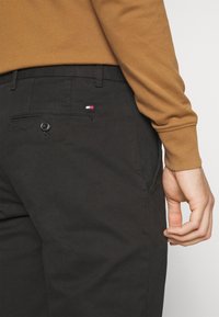 Pantaloni neri in cotone con una tasca posteriore con bottone e un piccolo logo. Chi indossa è vestito con una camicia a maniche lunghe marrone.