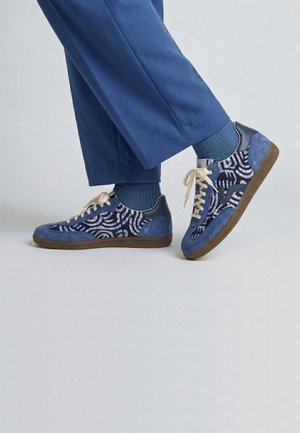 Personne portant des baskets bleues à motifs avec des lacets beige clair, des chaussettes bleues et un pantalon bleu sur un fond gris uni.