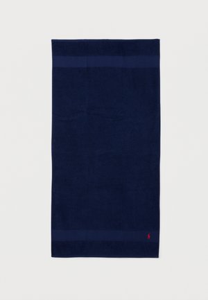 Toalla rectangular azul oscuro con una banda horizontal sutil cerca de los bordes y un pequeño logo rojo bordado en la esquina inferior.