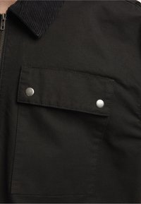 Czarna kurtka zapinana na zamek z kołnierzem z cordurowego materiału, przednia kieszeń na klapkę, srebrne zatrzaski oraz gładka, trwała tekstura materiału.