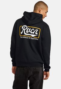 Black hooded sweatshirt featuring a bold white and yellow graphic text on the back, with "Rucn" and "The Balance Of Opposites." Textured fabric.