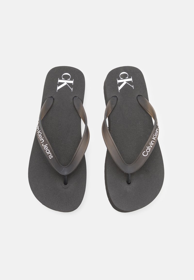 Mustat varvassandaalit, joissa on teksturoitu pohja ja kaksinkertaiset mustat hihnat. Varustettu valkoisella "ck"-logolla ja "Calvin Klein Jeans" -tekstillä. Suunniteltu rentoon käyttöön.