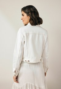 Chaqueta de mezclilla blanca con un diseño ajustado, detalles de botones en los puños y la parte trasera, y costuras visibles. Llevada sobre un vestido ligero y fluido.