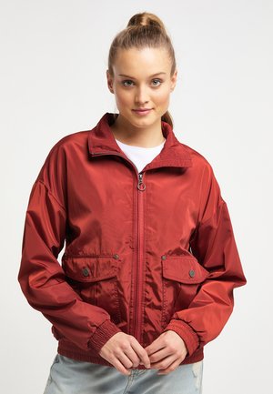 Giovane donna con capelli castano chiaro raccolti, indossa una giacca rossa con zip e tasche con pattina e jeans blu, in piedi davanti a uno sfondo uniforme.