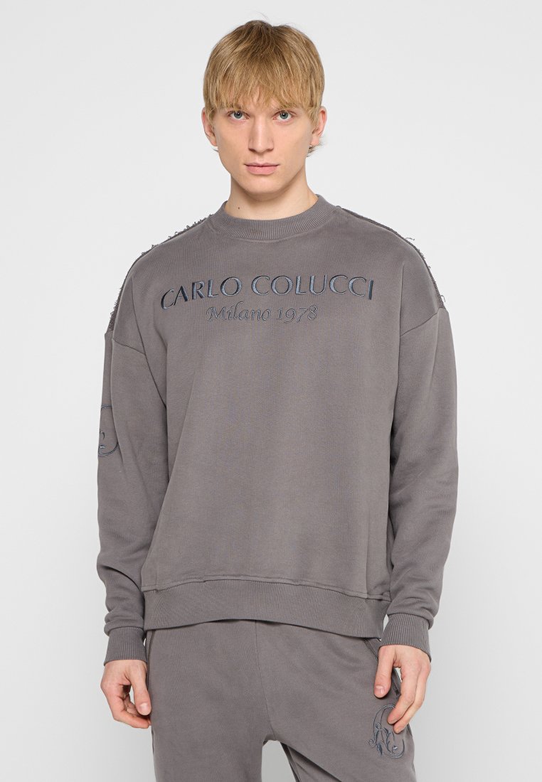 Junger Mann, der einen passenden grauen Sweatshirt und eine Hose trägt, auf dem Sweatshirt steht "Carlo Colucci Milano 1978" gestickt.
