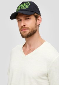 Schwarze Baseballmütze mit grünem "PURE JUICE"-Text und Zitronengrafik. Hellfarbiges T-Shirt mit V-Ausschnitt und weichem Textil. Neutraler Hintergrund.